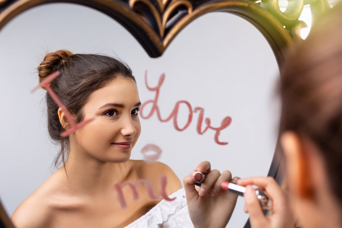 Portrait of a beautiful caucasian girl looking in the mirror and writing I love me near a heart, self-love